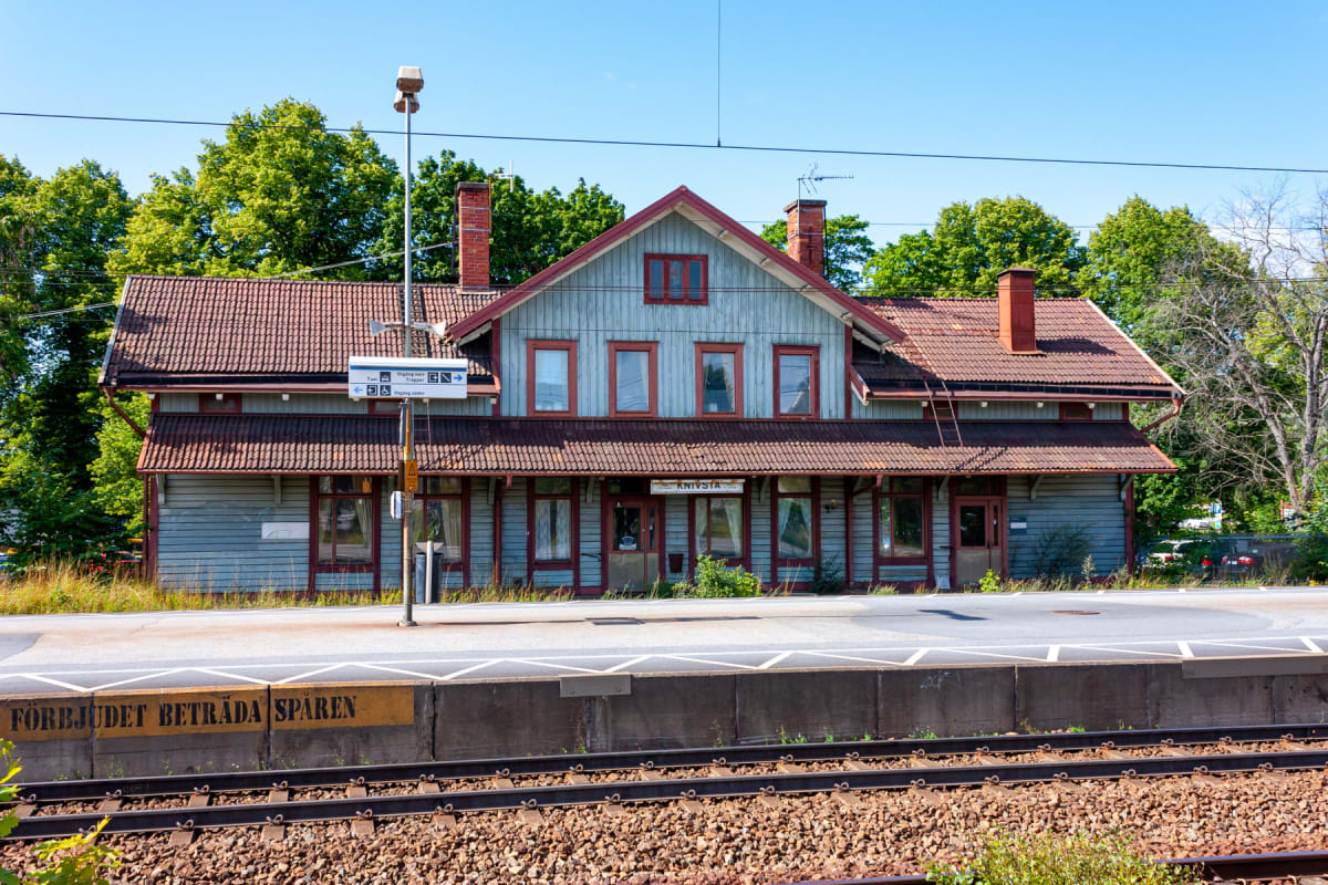 En bild som visar en ljusblå träbyggnad med rött tak och röda fönsterkarmar, troligen en stationsbyggnad, sedd från spåren på en tågstation. På byggnadens framsida står ordet "KNIVSTA" och även en skylt med hänvisningar syns.