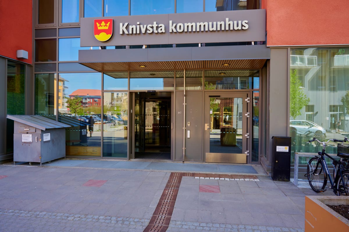 En fasadvy av entrén till Knivsta kommunhus, med skyltar som anger namnet. Byggnaden är i en orange färgton med stora glaspartier som reflekterar omgivningen. Utanför entrén står en cykel, en brevlåda och en återvinningslåda. Det syns också en person