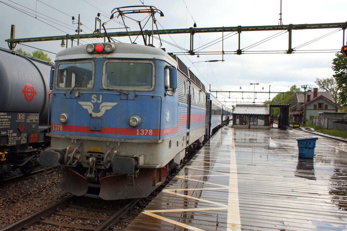 En blå och grå SJ-lokomotor med nummer 1378 står på ett järnvägsspår bredvid ett tankvagnståg. Till höger syns en regnvåt perrong med en liten station och i bakgrunden hus och träd under en molnig himmel. Kontaktledningar löper över spåren.