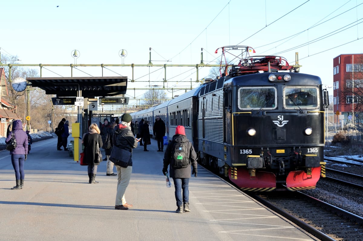 En svart SJ-loks front syns nära en perrong där människor står. Loket är numrerat "1365" och har röda snöplogar. På perrongen finns skyltar som visar "Spår 1 Uppsala" och "Spår 2 Märsta Stockholm". I bakgrunden syns ledningar för tågtrafik och ett rö