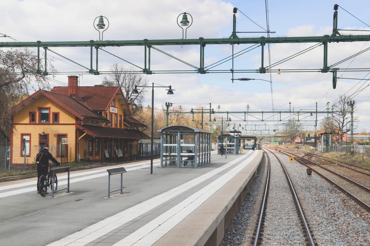 En bild av en järnvägsstation utomhus, föreställande en perrong med en man och en cykel. I bakgrunden är ett gult hus med rött tak och det är ledningar för tåg uppspända över spåren. Två glashus syns på perrongen och till höger är flera järnvägsspår.