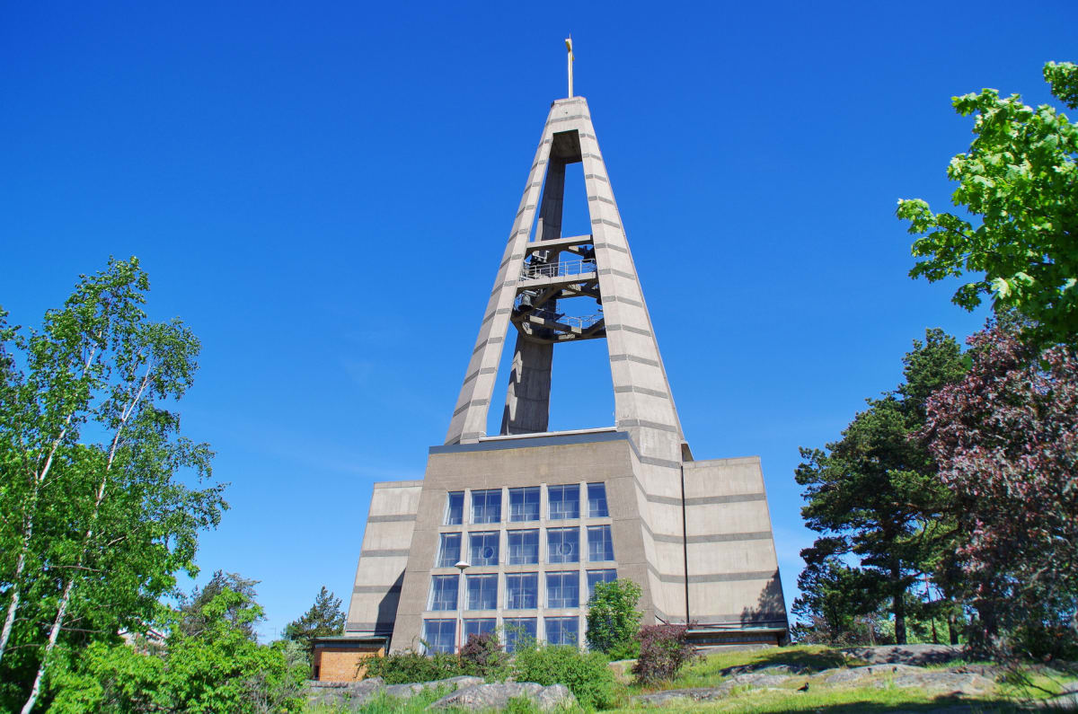 En modern kyrkobyggnad med en hög, spetsig klockstapel i betong under en klarblå himmel. Runt byggnaden växer lummiga gröna träd och buskar, med stenig terräng i förgrunden.