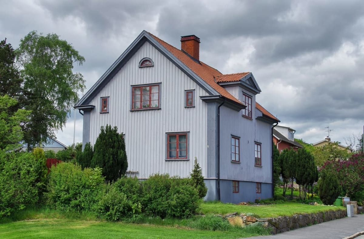 En ljusblå trävilla med rött tak och röda fönsterramar under en molnig himmel. Framför huset syns en gräsmatta, buskar och träd. Till höger finns en asfalterad yta och en stenmur.