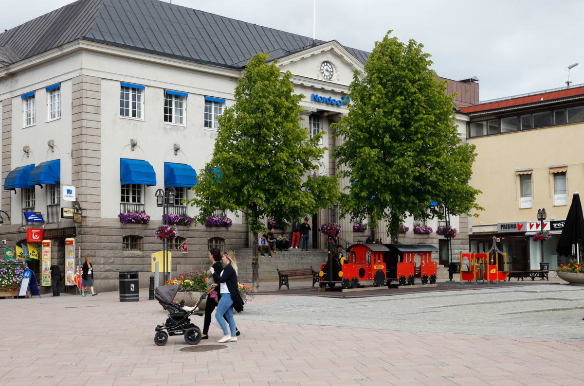 En bild som visar ett torg med byggnader i bakgrunden. Ett större vitt hus med blå markiser och skylten "Nordea" dominerar. Flera gröna träd, blomlådor och människor syns. På torget finns ett rött lektåg och en barnvagn. Flera personer rör sig på tor