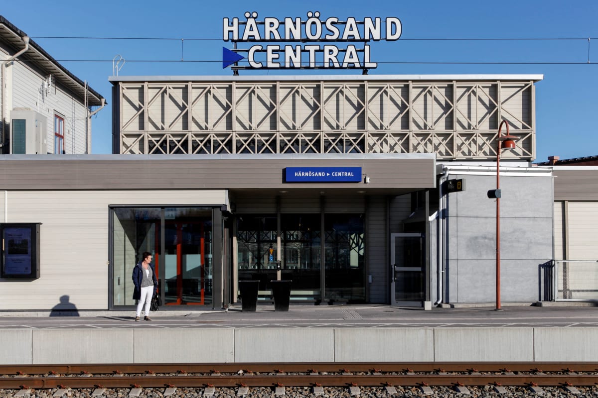 En kvinnlig person står på en tågplattform framför en byggnad med skylten "HÄRNÖSAND CENTRAL" under en klarblå himmel. Räls och grus syns i förgrunden.