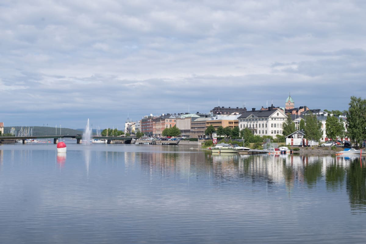 En bild av en stad vid vatten. I förgrunden syns vatten med reflekterande bebyggelse, i mellangrunden stadsbebyggelse, båtar och träd, och i bakgrunden en bro över vattnet och en trädklädd kulle. En fontän syns i vattnet.
