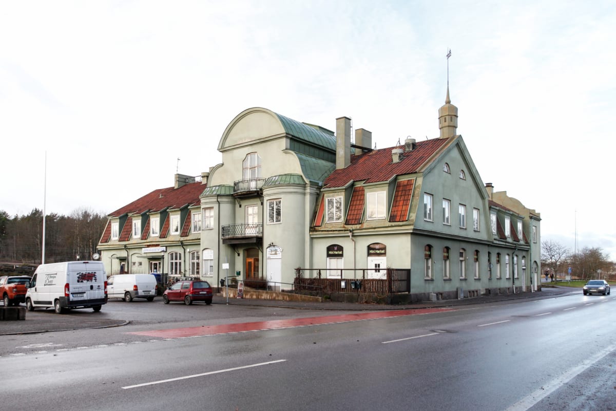 En stor, ljusgrön byggnad med rött tak och vindar står vid en väg. Flera bilar är parkerade utanför och en bil kör på vägen. Skyltar för 'Mini Market' och 'JÄRNVÄGSSTATION' syns. Väderleken ser ut att vara molnig.