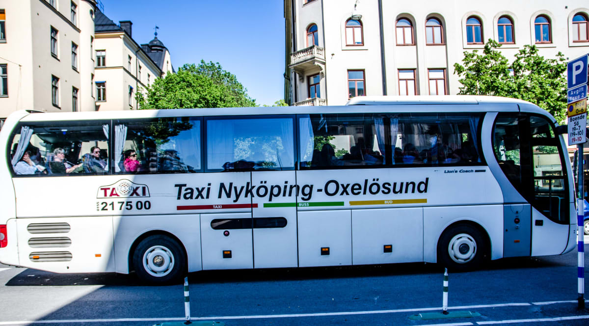 En vit buss med texten "Taxi Nyköping-Oxelösund" står parkerad. Flera personer syns inuti bussen. I bakgrunden syns ljusa byggnader och gröna träd under en blå himmel. En parkeringsskylt med text på svenska syns till höger om bussen.