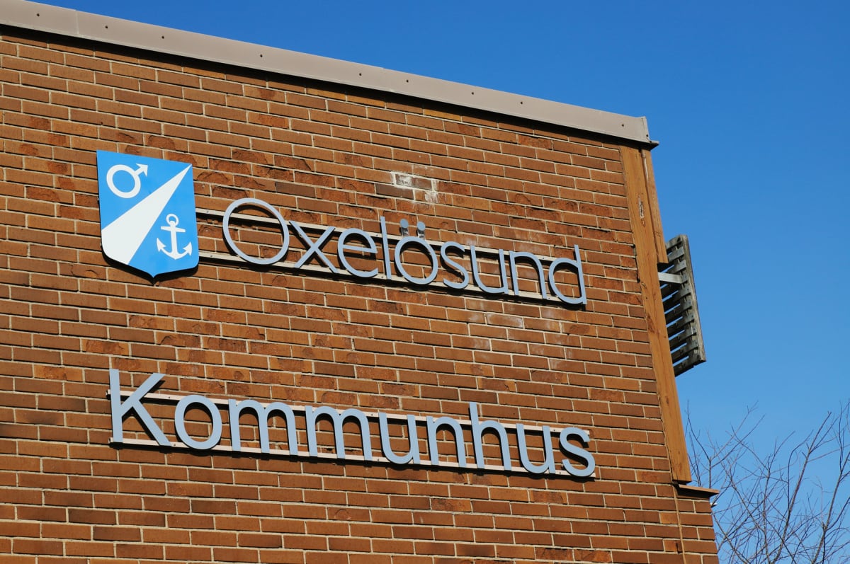 En fasad av rött tegel med en plakett och texten "Oxelösund" och "Kommunhus" i silverfärgade bokstäver. Plaketten är blå och vit med en symbol som liknar en manssymbol och ett ankare. Bakgrunden är en klarblå himmel.