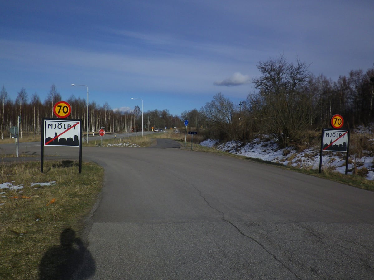 En vägkorsning med asfaltvägar, vägskyltar och snöfläckar på sidorna. I bakgrunden syns skog med lövfällande träd och en klarblå himmel med några moln. En trafikerad väg sträcker sig framåt med fordon synliga längre bort.