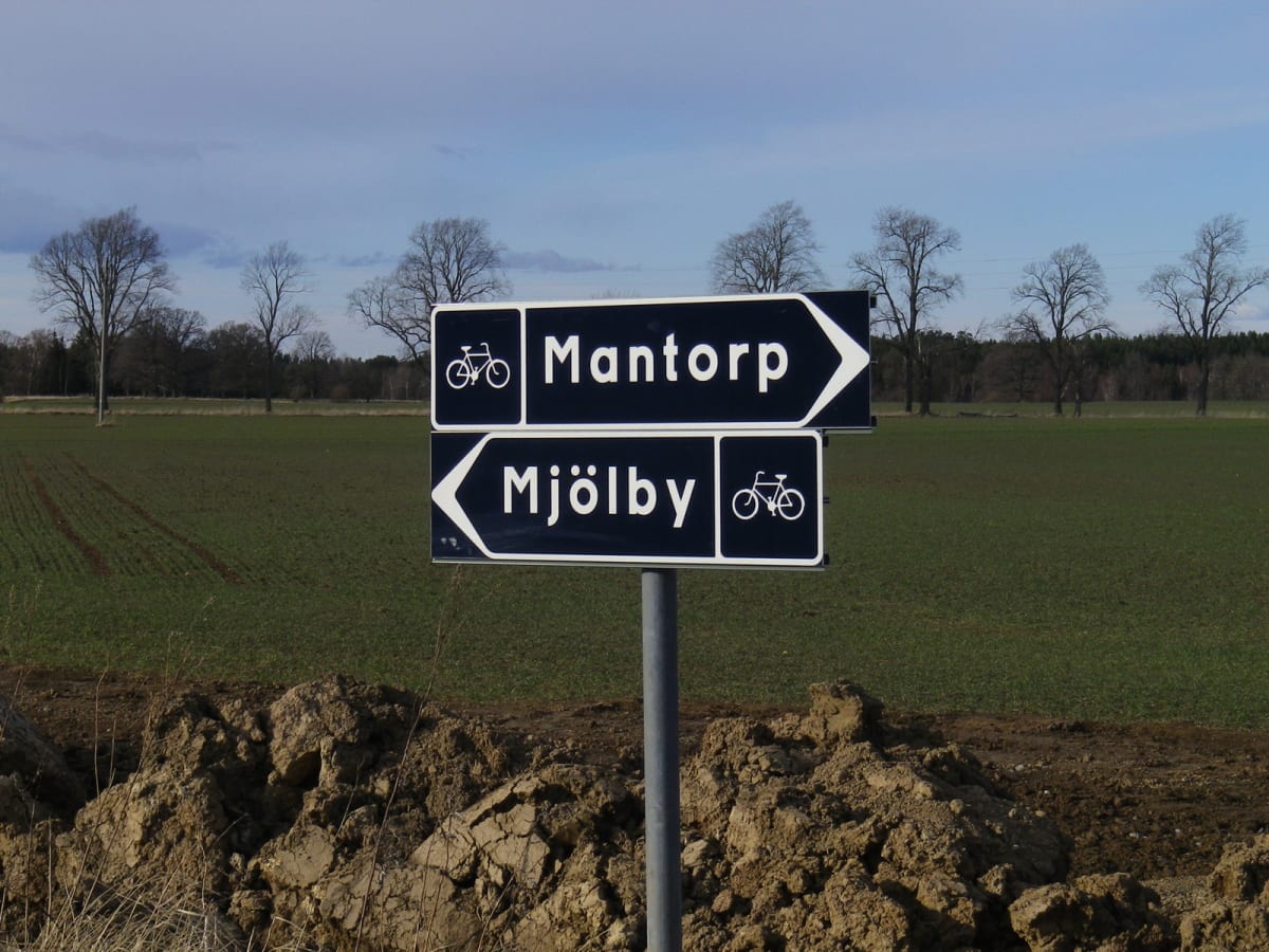 En svart och vit vägskylt på en grå stolpe, med vita pilar och texten "Mantorp" samt "Mjölby", med symboler för cykel. Skylten står i ett grönt fält med träd i bakgrunden under en blå himmel med moln. I förgrunden syns en jordhög.