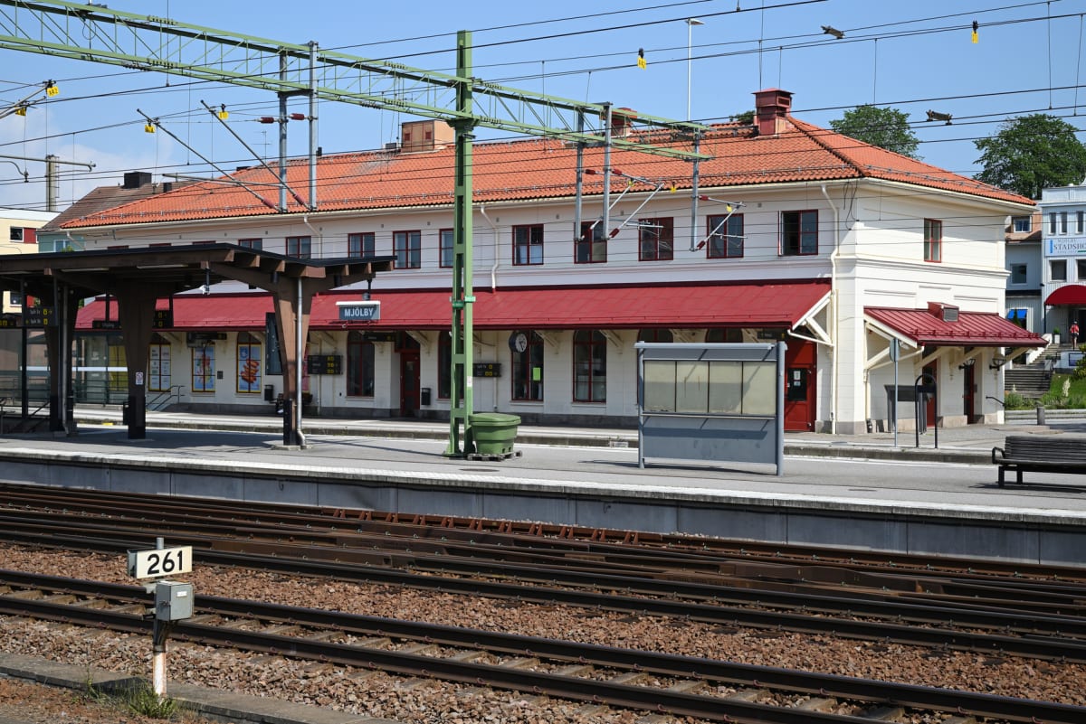 En bild som visar en järnvägsstation med ett stort, ljust byggnad med rött tak. I förgrunden syns tågspår med makadam och en skylt märkt "261". Ovanför spåren finns ett stort nätverk av kontaktledningar och stolpar. En stationsplattform ligger intill