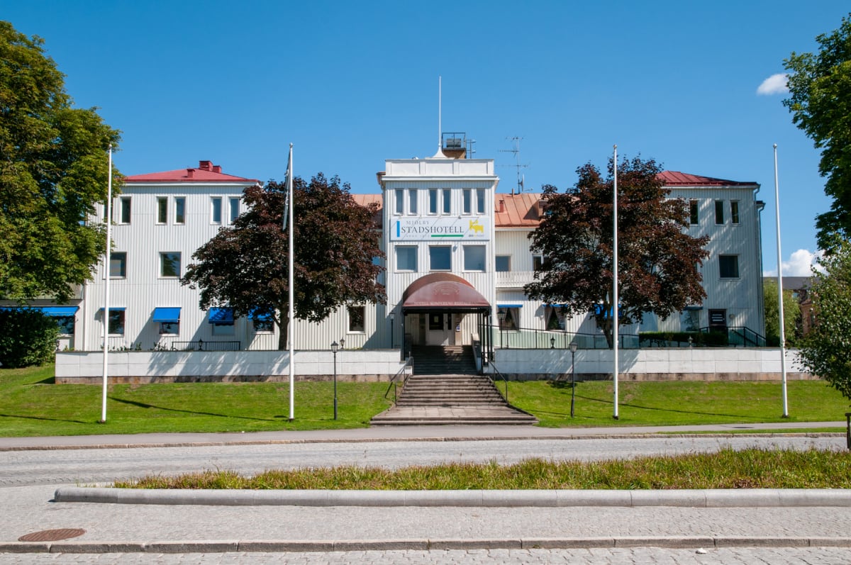 En stor, vit byggnad med rött tak och blå markiser på vissa fönster. Två flaggstänger står framför byggnaden som har en stentrappa upp till entrén. Gräsmatta framför byggnaden och träd på sidorna.