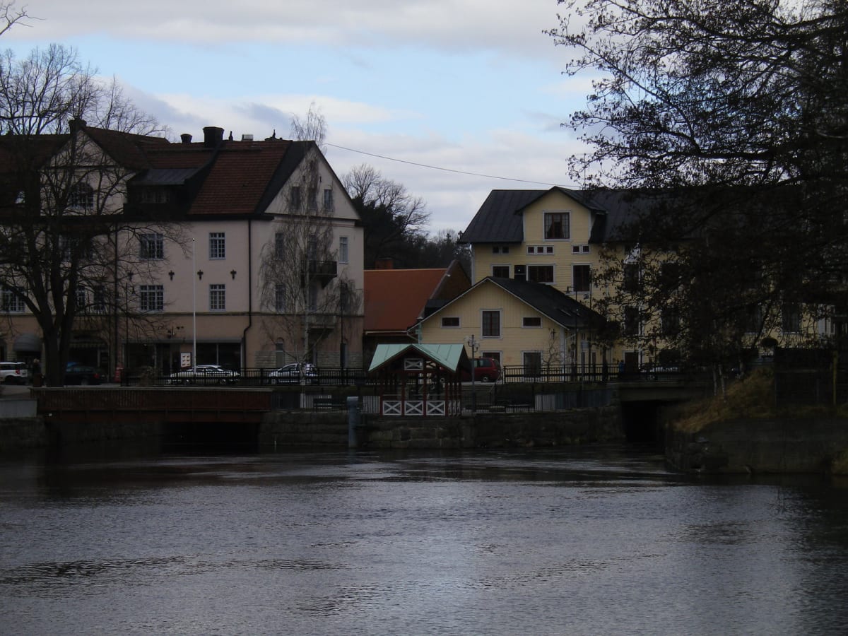 En bild som visar en flod eller å med hus på båda sidor av vattnet. Flera broar korsar vattnet. Träd med kala grenar står längs stranden och framför byggnaderna. Himlen är delvis mulen med ljusblå fläckar. Flera fordon syns bland husen.