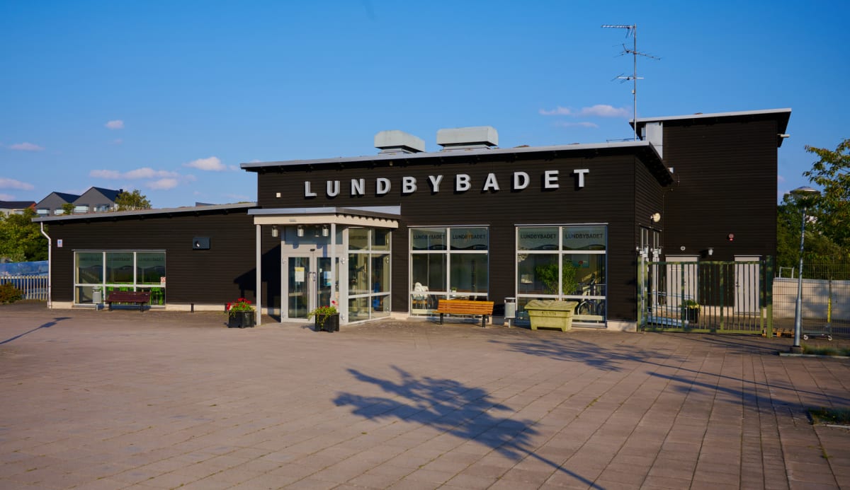En modern byggnad med mörka fasader och vita bokstäver som stavar "LUNDBYBADET". Byggnaden har stora fönsterpartier och en entré med glasdörrar. Utanför finns en asfalterad yta med bänkar, växtlighet och en grön soptunna. Skuggor från träd faller på