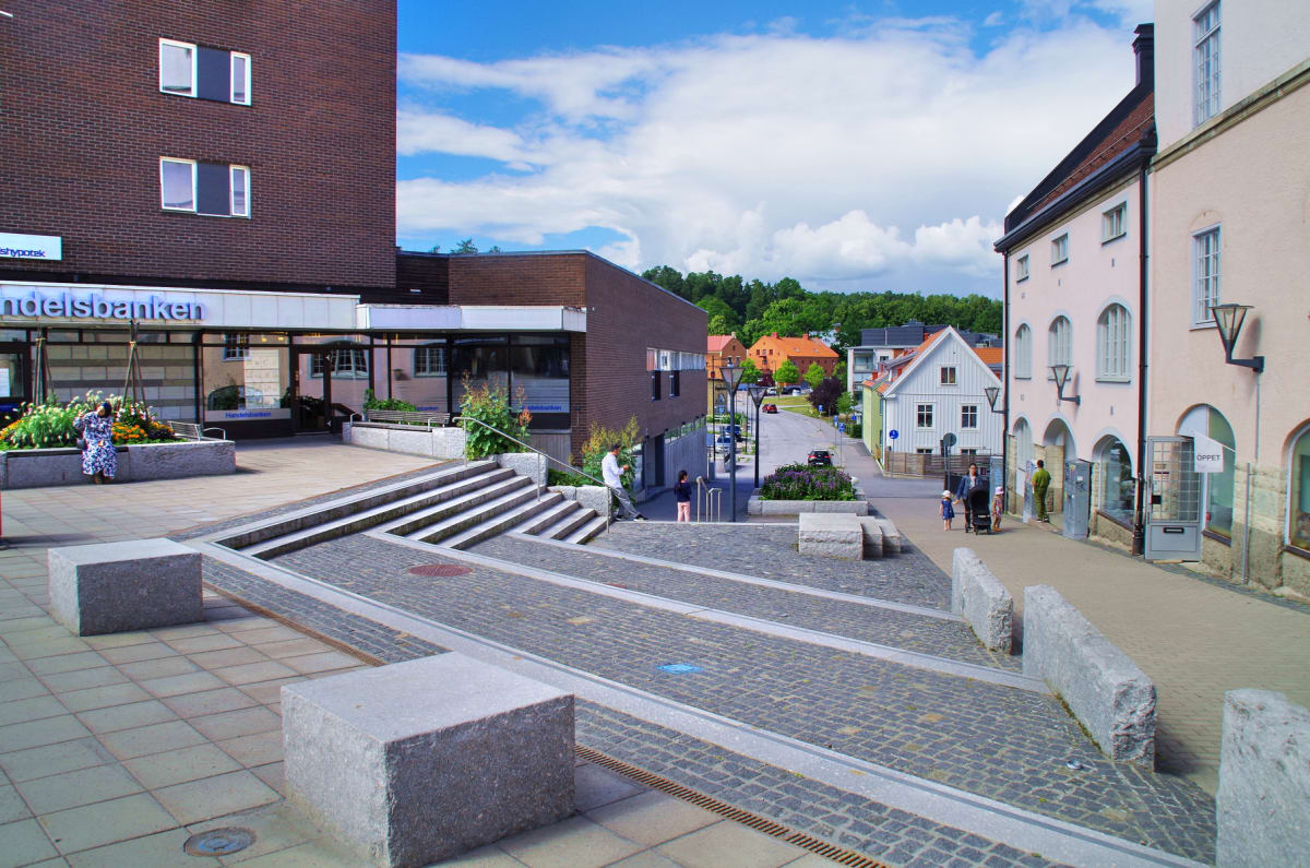 En gatsvy över ett mindre samhälle under dagtid med flera byggnader, en väg i bakgrunden och träd. På torget i förgrunden syns stenbeläggning, trappor och planteringar. Flera människor är synliga, däribland en kvinna vid en blomrabatt och en familj p