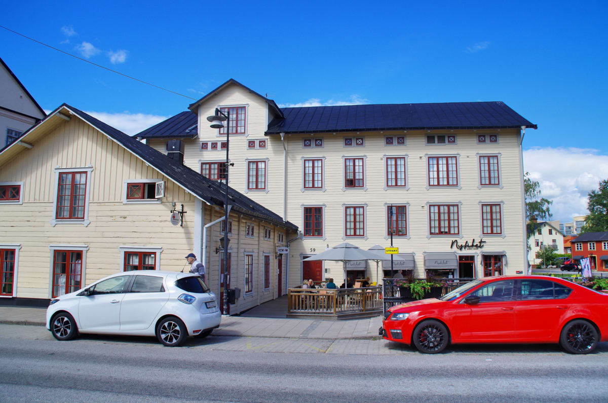 En gata med parkerade bilar och byggnader i bakgrunden. Två bilar, en vit och en röd, står parkerade vid trottoarkanten. Byggnaderna har ljusa fasader och mörka tak, med flera fönster. En uteplats med sittande personer syns framför en av byggnaderna