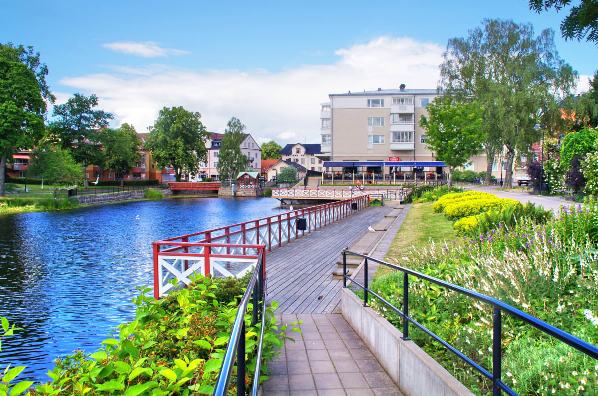 En bild visar en vattendrag med en lång träbrygga längs stranden, med en röd och vit räcke. På bryggan syns två måsar. På andra sidan vattnet finns bostadshus och grönska, samt en röd bro. På bryggans sida finns en gräsmatta med blommor och träd.
