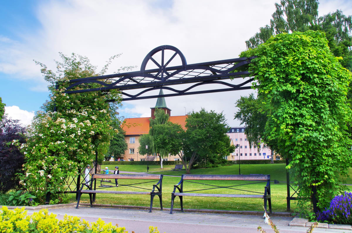 En översiktsbild av en park med två parkbänkar i förgrunden, omgivna av grönska och blommor. I bakgrunden syns en byggnad med orange tak och en grön spira, samt andra byggnader och träd. En metallkonstruktion täckt av klätterväxter sträcker sig över