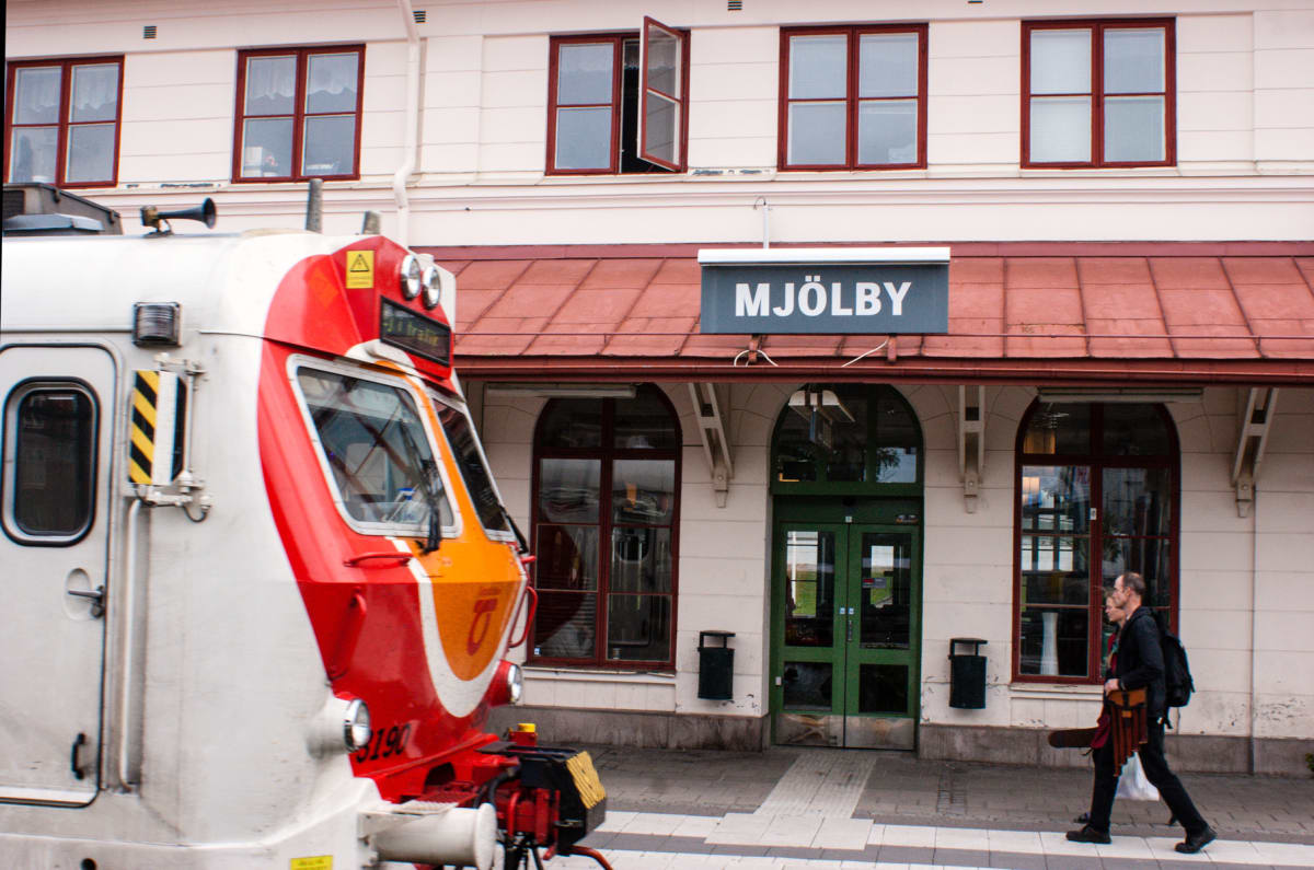 Bilden visar ett tåg som passerar en byggnad med en skylt där det står "MJÖLBY". En person går på trottoaren vid byggnaden. Byggnaden har flera fönster och dörrar samt ett rött tak. Högst upp på byggnaden finns tre våningar med fönster. Ett av fönstr