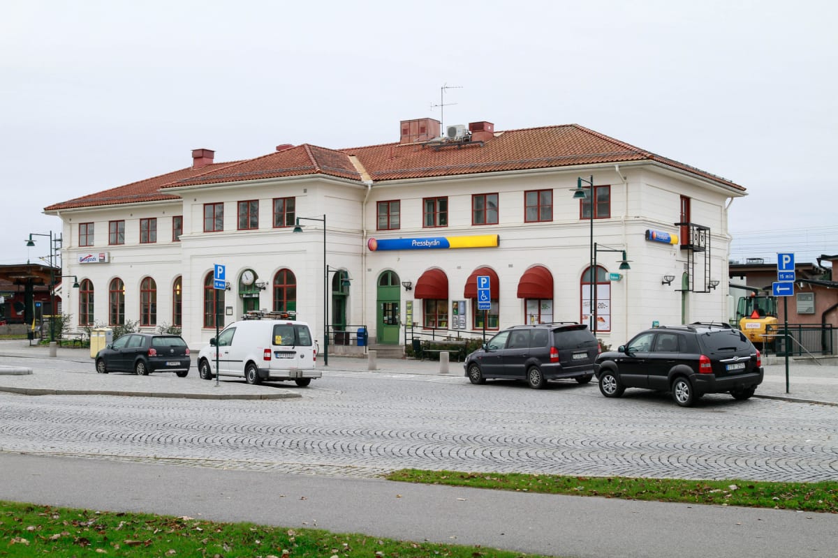 En bild som visar en ljus byggnad med rött tak framifrån, med bilar parkerade på kullerstensbelagd mark framför. Skyltar med texten "Pressbyrån" och parkeringsskyltar syns tydligt. En grön gräsremsa med vissna löv kan ses i förgrunden.
