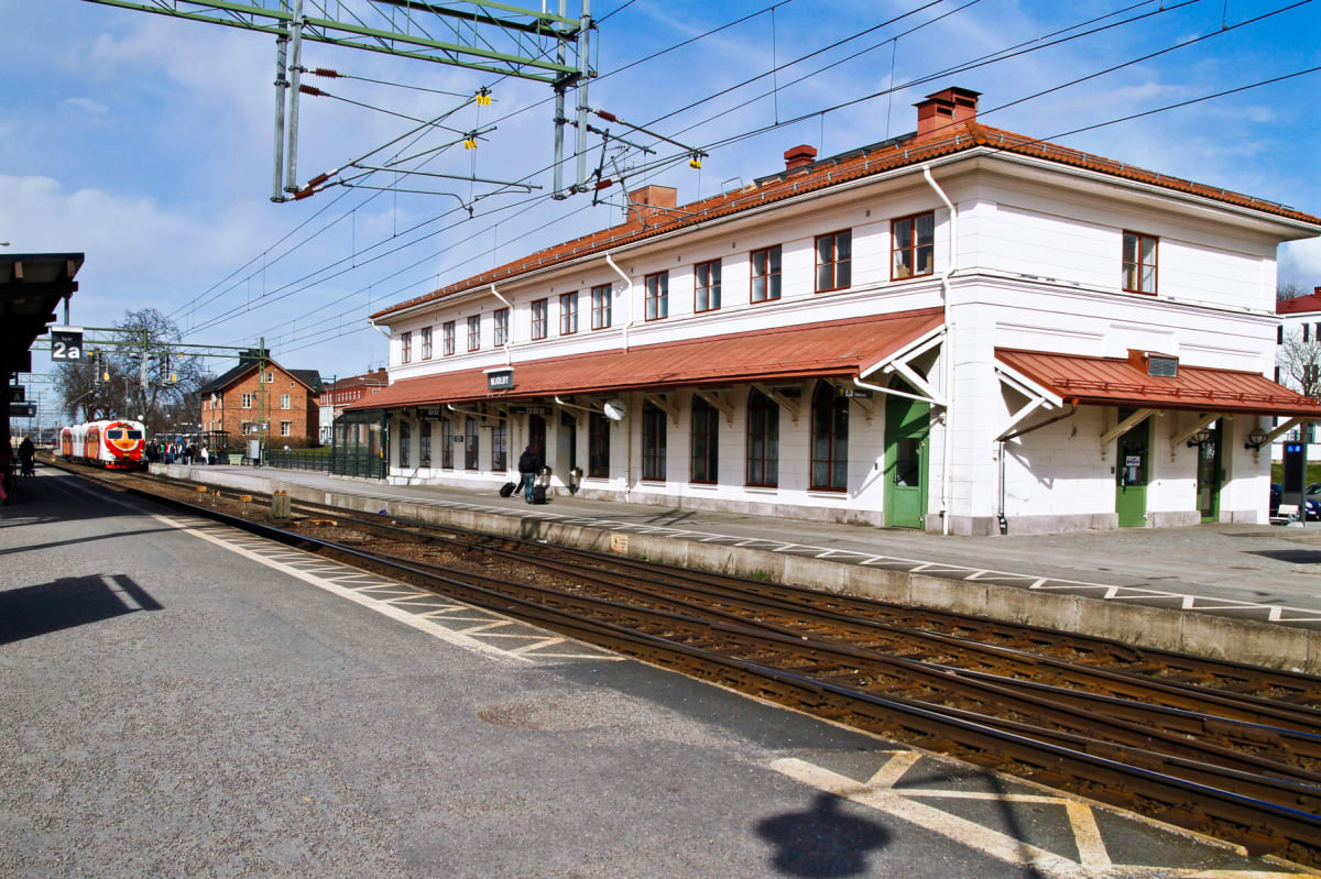 En bild av en tågstation med ett vitt stationshus med rött tak och en plattform. Ett orange och vitt tåg står vid en spår. Flera personer syns på plattformen och en person med resväska nära stationshuset. Blå himmel med några moln.