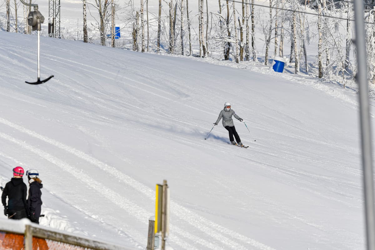 En person åker skidor nedför en snötäckt backe. Till vänster i bakgrunden syns ett ankarlift, och i bakgrunden finns snötäckta träd. Ytterligare två personer med hjälmar syns i nedre vänstra hörnet, delvis avskurna av bildkanten.