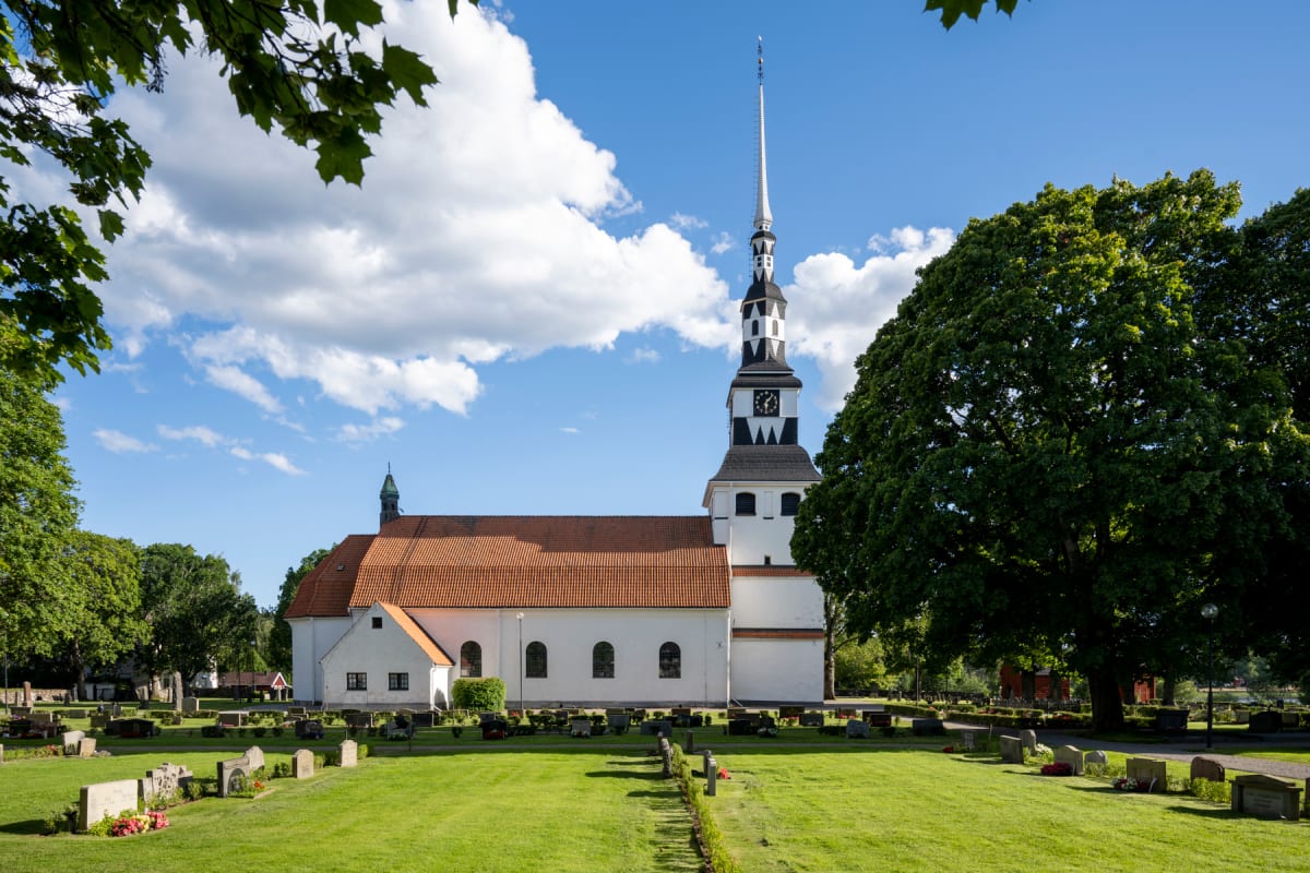 En kyrka med vit puts och tegeltak syns under en blå himmel med moln. Till vänster och höger om kyrkan skymtar trädkronor. Framför kyrkan ligger en grön gräsmatta med ett flertal gravstenar. En stor, grönskande trädtopp dominerar bildens högra sida.