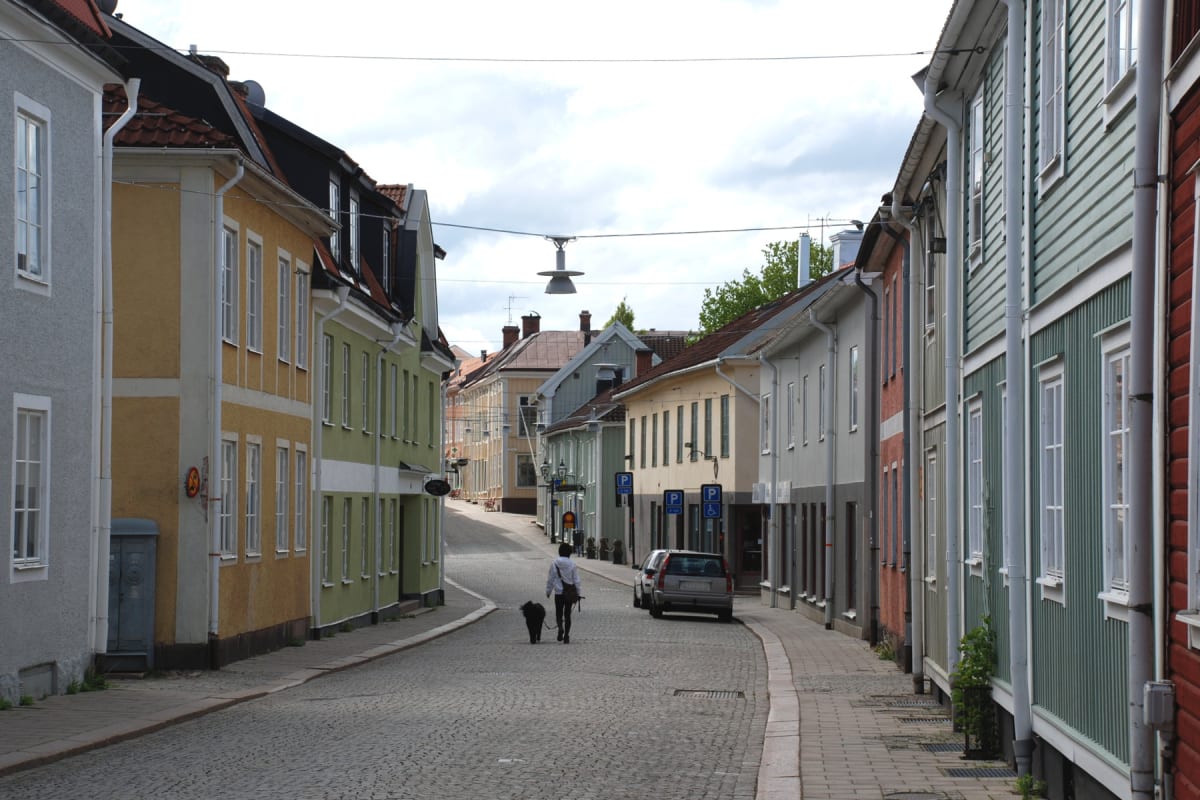 En gata kantad av flervåningshus med fönster. En person går med en hund på kullerstenen, och en parkerad bil står vid sidan av gatan. Ljusa moln syns på himlen ovanför. Husen har varierande färger, bland annat gult, grönt, rött och grått. Gatubelysni