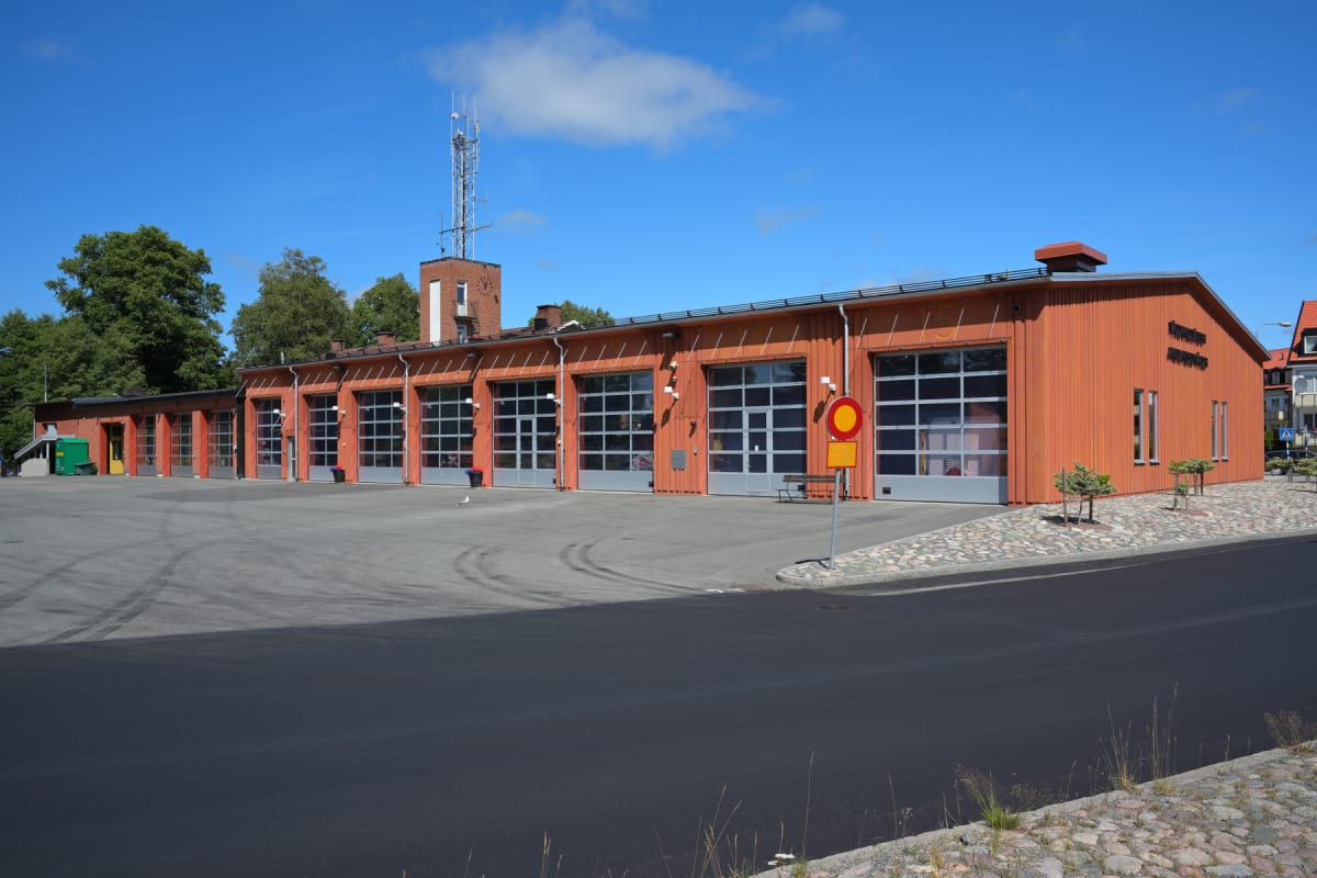 En modern brandstation med sju portar står under en klarblå himmel med några moln. Byggnadens fasad är orangefärgad och har en tegelbyggnad med en antennmast på taket i bakgrunden. Framför portarna finns en asfalterad yta, delvis med däckspår.