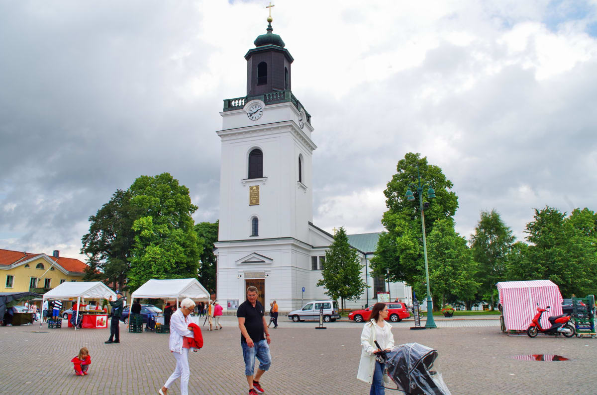 En stor öppen plats med en vit kyrka med klocktorn i mitten. Flera personer, inklusive ett barn, går över platsen. Två marknadsstånd med vita tältdukar och ett med röd- och vitrandig tältduk står uppställda. Gröna träd omger platsen under en molnig h