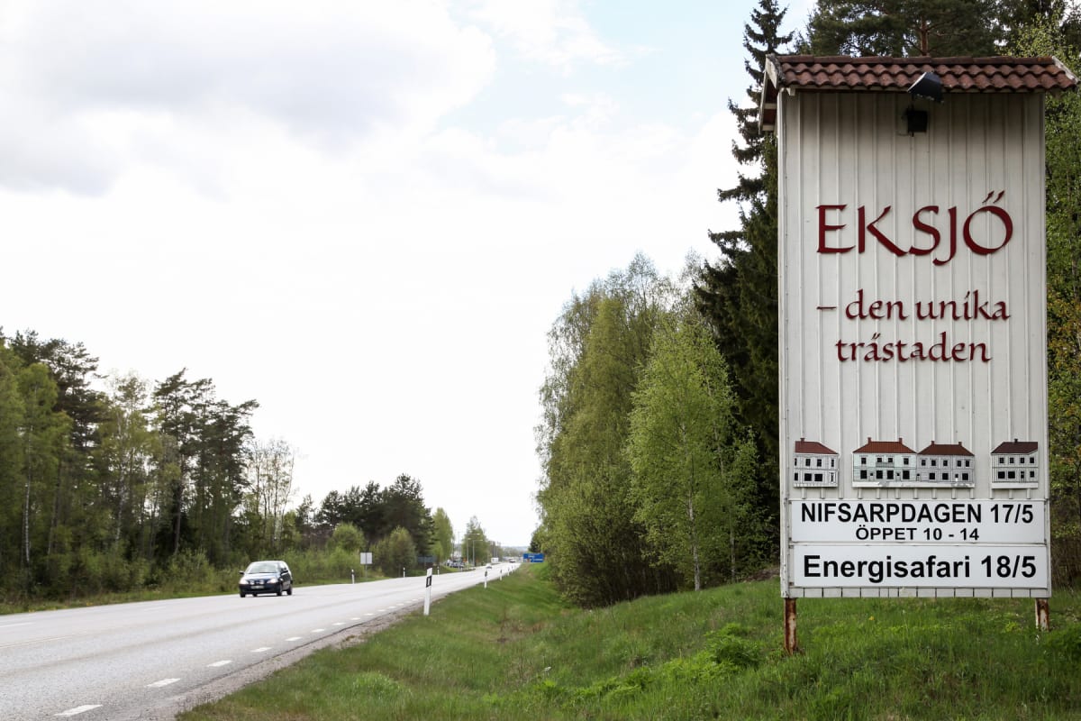 En stor skylt med texten "EKSJÖ - den unika trästaden" och illustrationer av hus står vid sidan av en landsväg. En bil kör på vägen som kantas av träd och gräs. Himlen är delvis molnig.