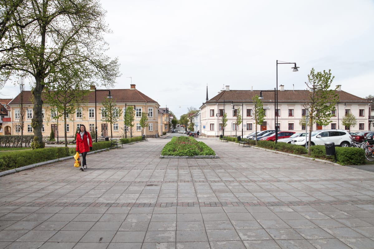 En individ går på en stensatt gångväg, med träd, buskar och blommor intill. Bakom personen finns byggnader med flera fönster och takpannor. En parkeringsplats med bilar syns till höger, och en väg sträcker sig i bakgrunden.