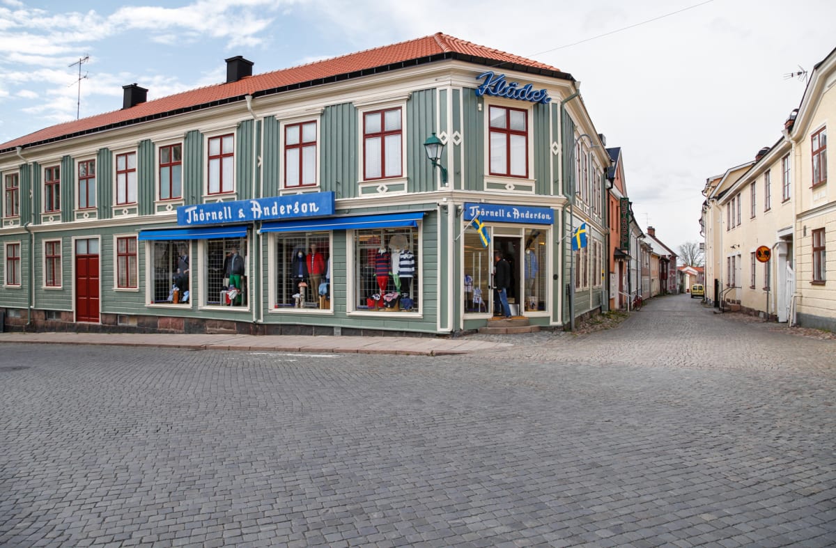 En gathörna med en butik med texten "Thörnell & Andersson" på fasaden och "Kläder" på taket. Butiken saluför kläder, utställda i skyltfönstren. Gatan framför är belagd med kullersten. Längs gatan i höger syns ytterligare byggnader och parkerad bil.