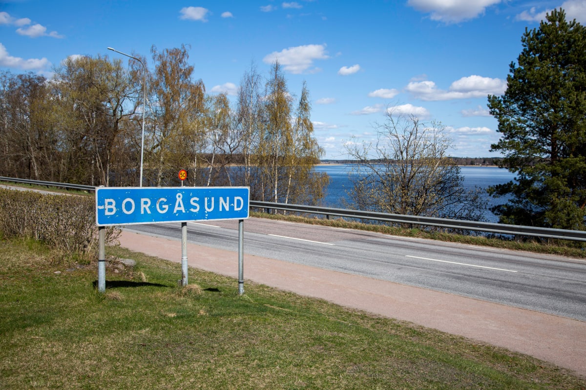 En bild tagen utomhus under dagtid, som visar en blå ortnamnsskylt med texten "BORGÅSUND" vid sidan av en asfalterad väg. I bakgrunden syns en stor vattenyta omgiven av träd och en blå himmel med moln.