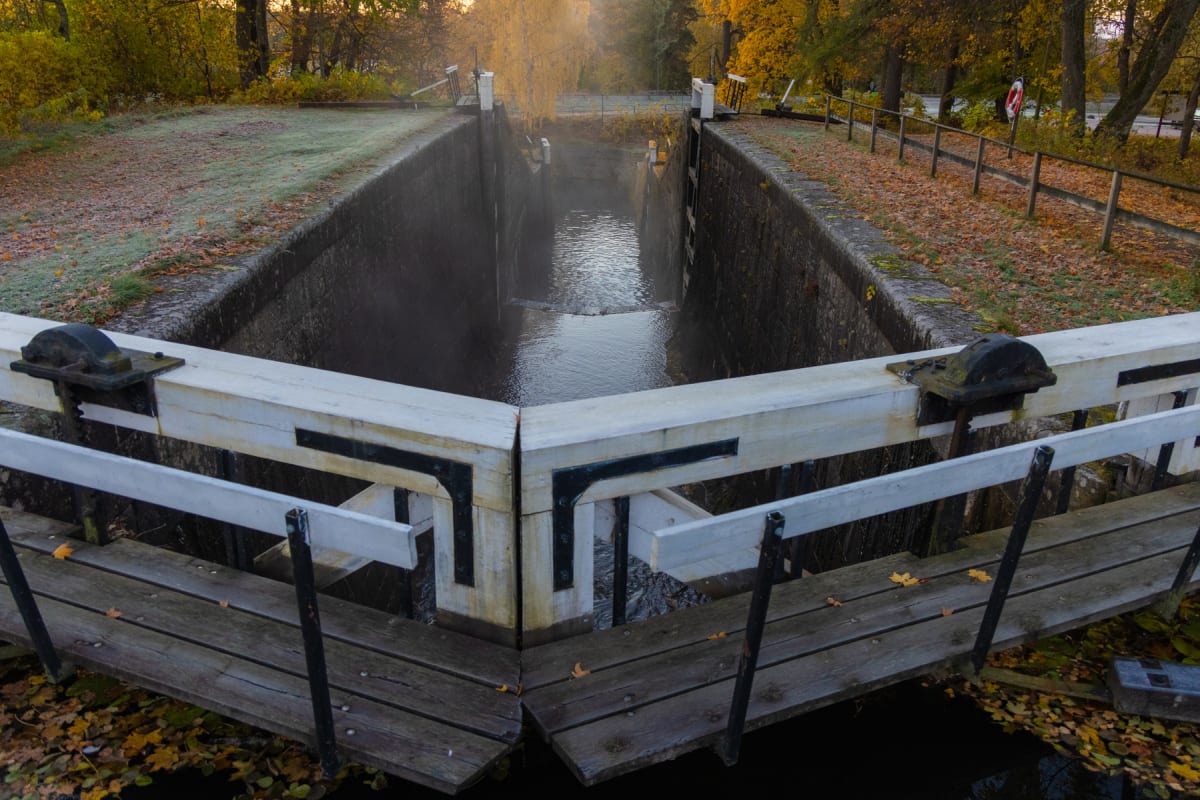 En bild som visar en slusskanal kantad av höstlövsfärgade träd. Vattnet i slussen är mörkt med en antydan till dimma, och landytorna ovanför slusskanalen är täckta av höstlösa träd och ett tunt lager frost. En livboj syns på höger sida.