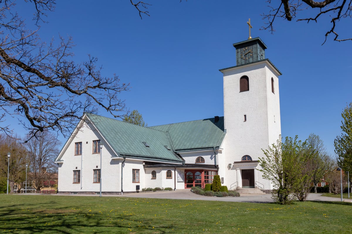 En vit byggnad med grönmossigt tak och ett klocktorn med ett guldkors på toppen står under en klarblå himmel. Gräsmattor omger byggnaden och träd med både nakna grenar och små, gröna löv syns i förgrunden och bakgrunden.