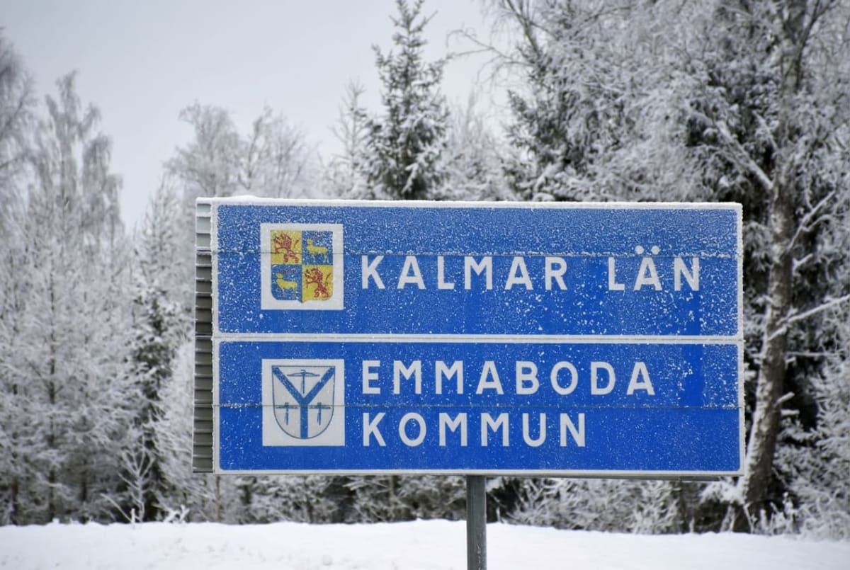 En blå skylt med vita bokstäver där det står "KALMAR LÄN" och "EMMABODA KOMMUN", med respektive vapensköldar, står täckt av snö och frost. I bakgrunden syns snötäckta träd under en grå vinterhimmel.