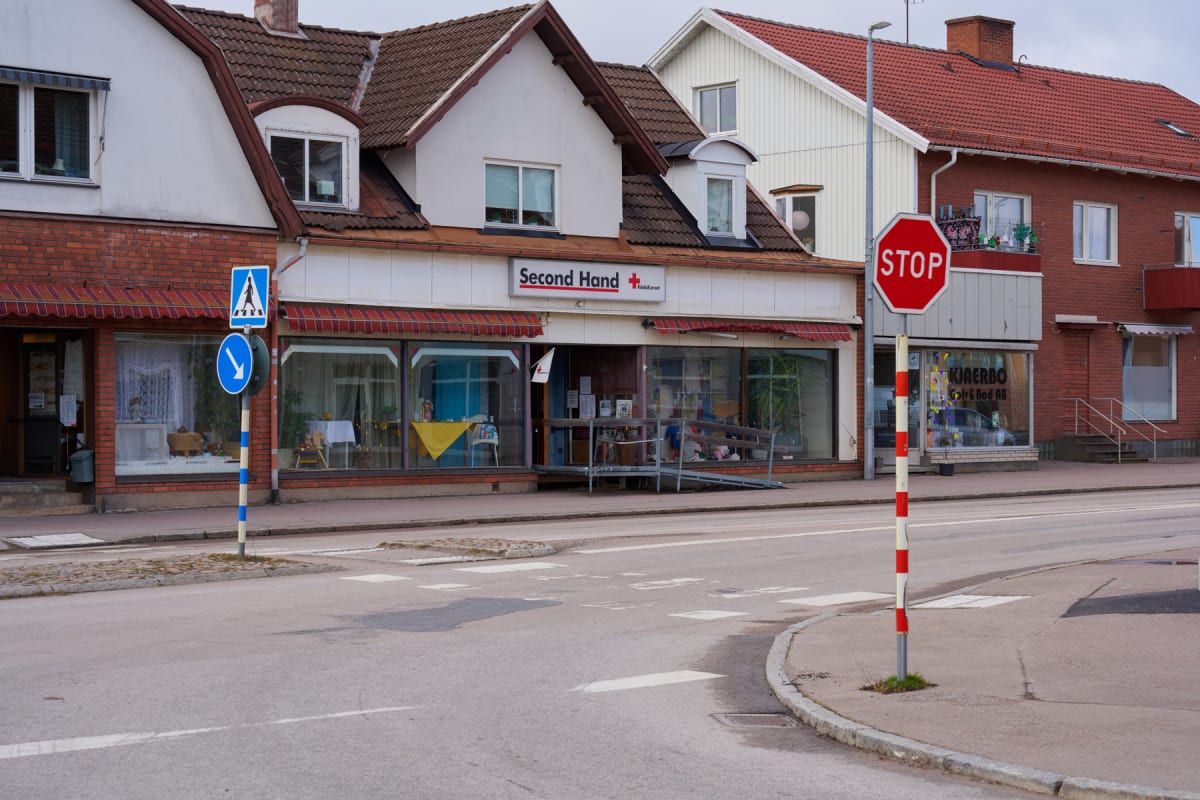 En ganska bred bild av en gata med byggnader i bakgrunden. Ett trafikljus, en stoppskylt och en övergångsskylt syns tydligt. Byggnaderna har tegelväggar på markplan och putsfasader på de övre våningarna. En butik med texten "Second Hand" och Röda Kor
