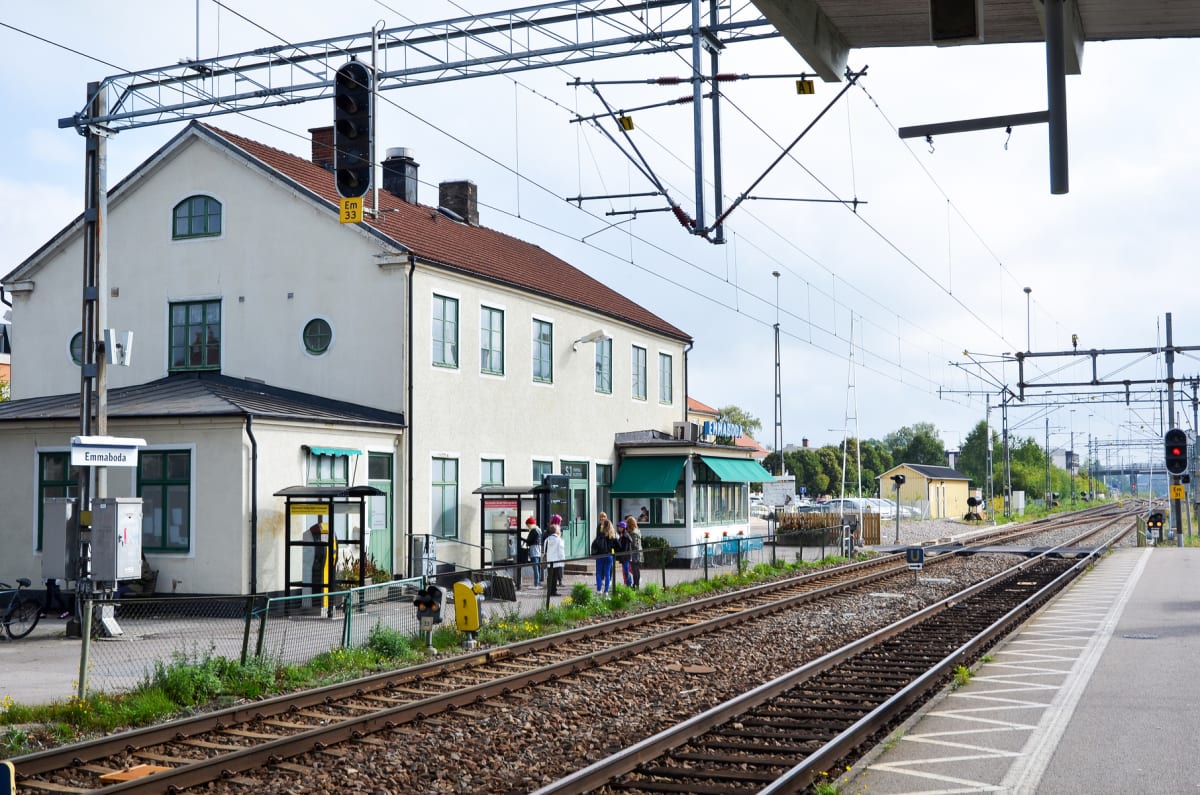 En bild som visar en järnvägsstation med ett beige stationshus med brunt tegeltak och gröna fönsterluckor. Framför stationshuset står flera människor och ett gult tåg verkar invänta passagerare. Två järnvägsspår sträcker sig mot horisonten. En upplys