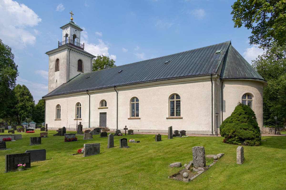 En beige stenkyrka med mörkblått tak och en hög klockstapel med gyllene kors, omgiven av en grön gräsmatta med gravstenar och träd, under en klarblå himmel med moln.