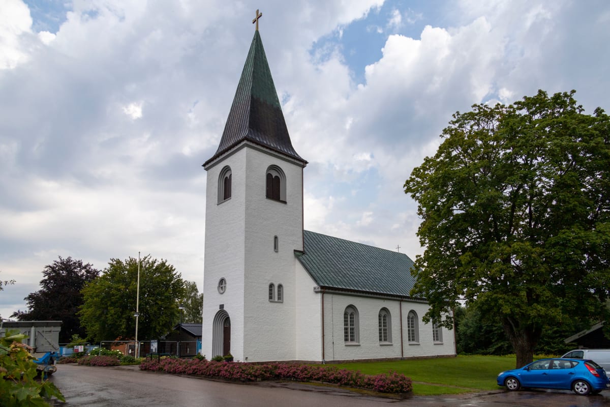 En vit kyrka med ett spetsigt mörkt tak och ett kors på toppen, står under en molnig himmel. Framför kyrkobyggnaden syns en gräsmatta med ett blomsterland och en asfalterad väg. En blå bil står parkerad på höger sida om kyrkan, bredvid ett stort träd