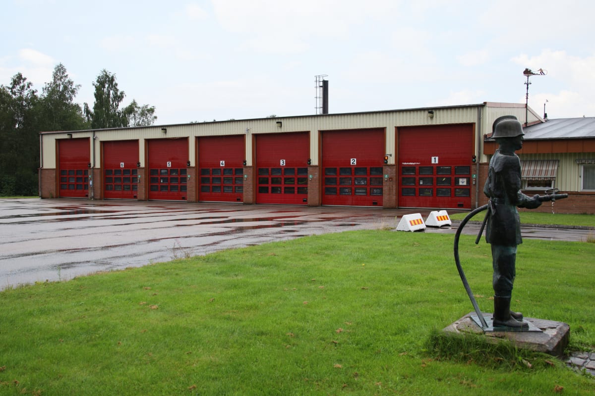 En bild som visar en brandstation med röda portar, gräsmatta och en staty av en brandman. Marken framför stationen är blöt med vattenpölar. Ett träd syns i bakgrunden till vänster och en skorsten på taket.