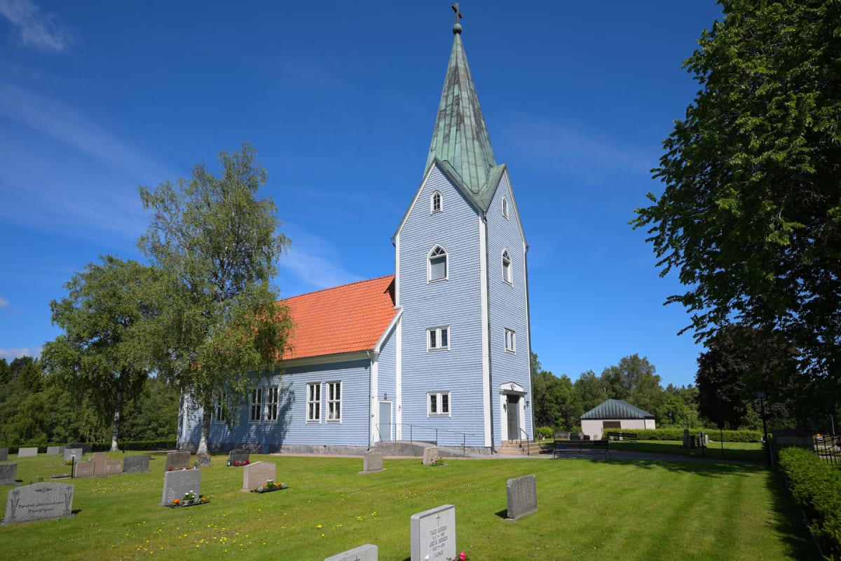 En kyrka med blå träpanel och tegeltak syns från en kyrkogård med gräsmattor, träd och gravstenar under en blå himmel. Kyrktornet har spetsigt tak och ett kors överst. En mindre byggnad med mörkgrönt tak skymtar i bakgrunden.