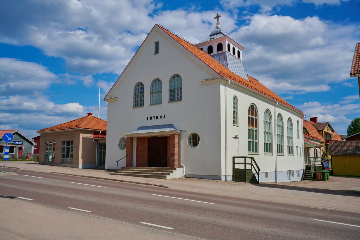 En vit kyrkobyggnad med orange tak står vid en väg under en ljusblå himmel med moln. Två vita linjer markerar vägen framför. Byggnaden har "SMYRNA" skrivet på framsidan och ett kors på toppen.