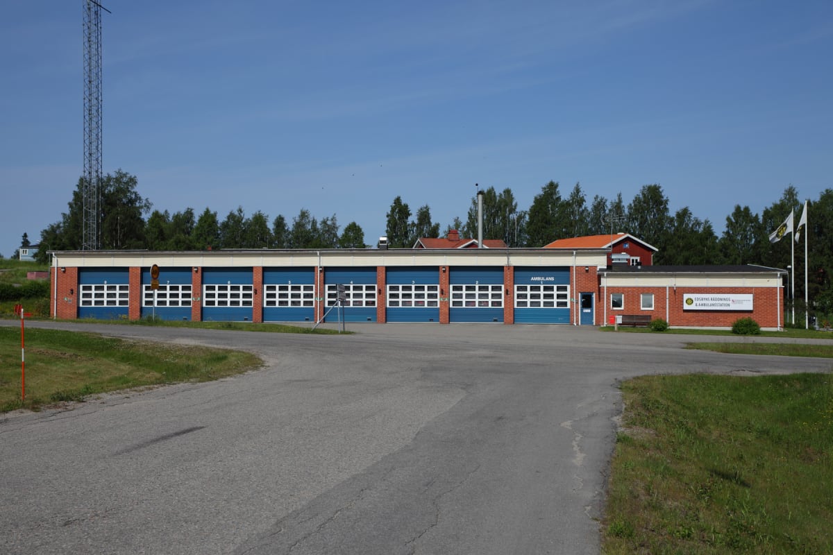 En röd tegelbyggnad med blå portar, tydligt markerad som en räddnings- och ambulansstation, omgiven av träd och gräs under en klarblå himmel. Ett asfalterat område syns framför byggnaden, och en mast reser sig i bakgrunden.