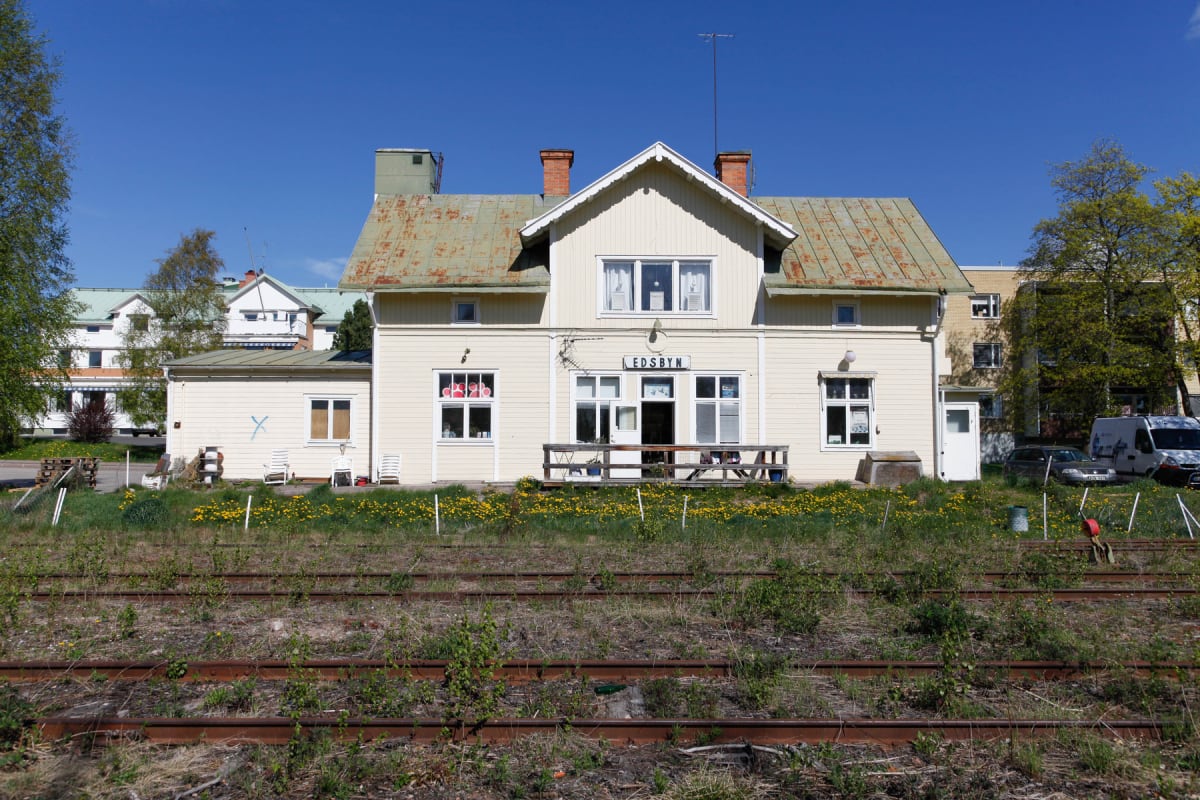 En gulbeige byggnad med namnet "EDSBYN" ovanför entrén. Framför byggnaden finns en altan med stolar och bord, samt ett område med gräs och gula blommor. I förgrunden syns järnvägsräls med gräs och vegetation. I bakgrunden syns andra byggnader och ett