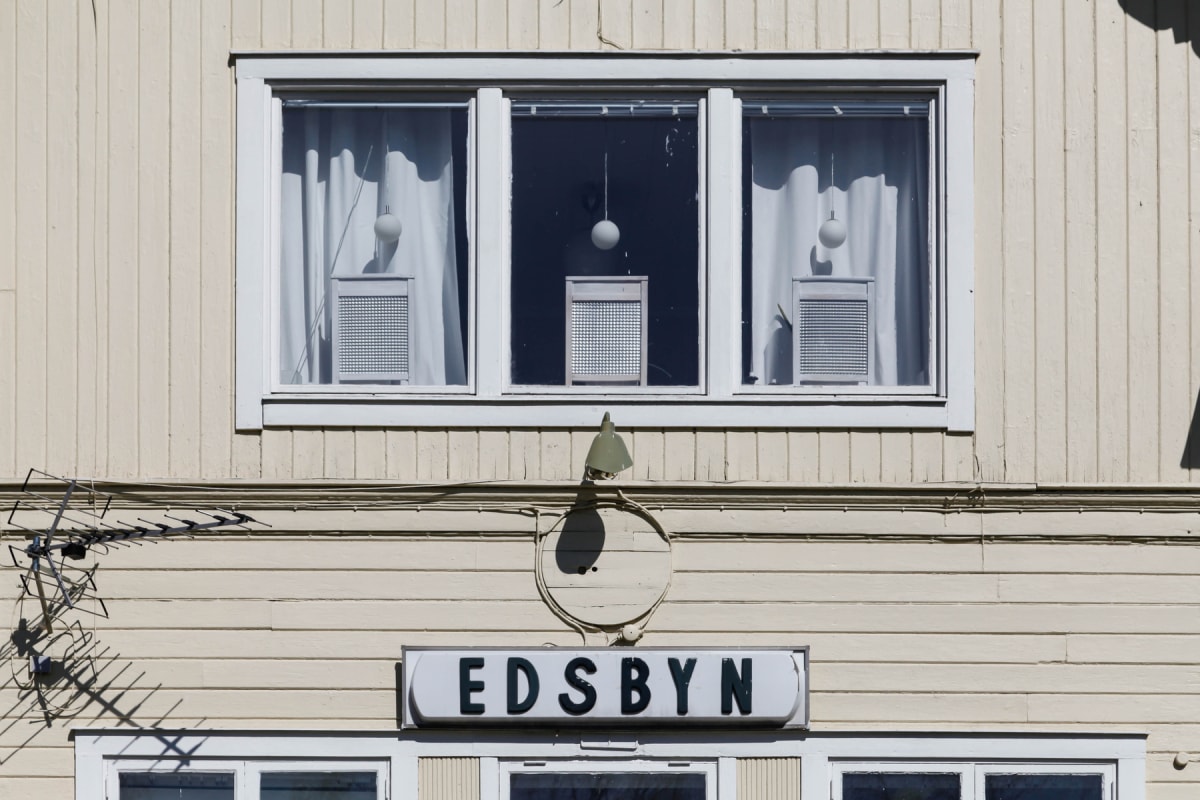 En fasad med tre fönster där vita gardiner och lampor syns inuti. Under fönstren finns en vägglampa och en skylt med texten ”EDSBYN”. En tv-antenn är monterad till vänster om skylten.