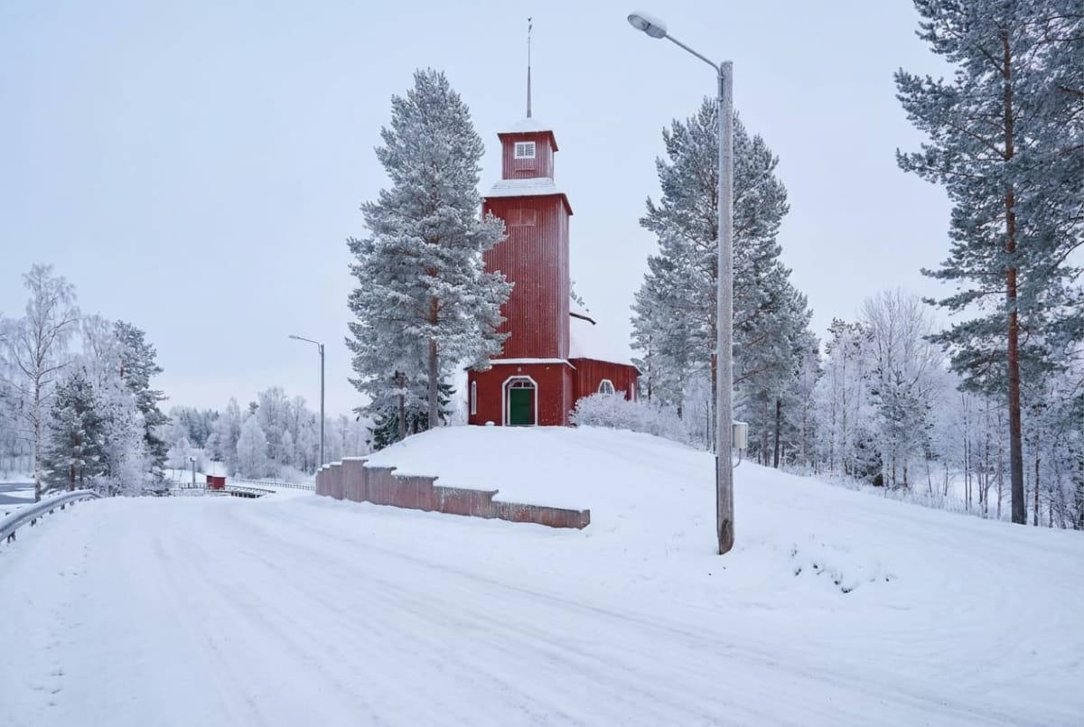 En röd träbyggnad med ett torn och en grön dörr, omgiven av snötäckta träd och mark. En snötäckt väg leder förbi byggnaden. Belysningsstolpar syns i förgrunden och bakgrunden. Träden är täckta av rimfrost eller snö.