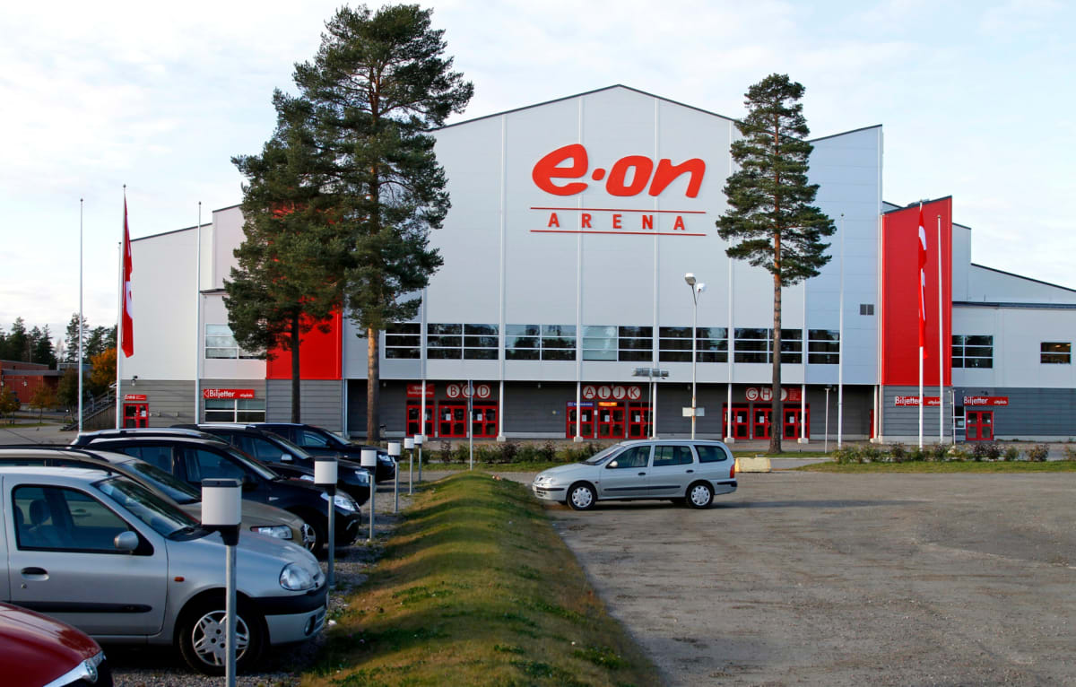 En stor, vit byggnad med texten "E.ON ARENA" i rött på fasaden. Framför byggnaden finns en parkeringsplats med flera parkerade personbilar. Två höga tallar står framför byggnaden och en röd flagga hänger från en flaggstång till vänster.